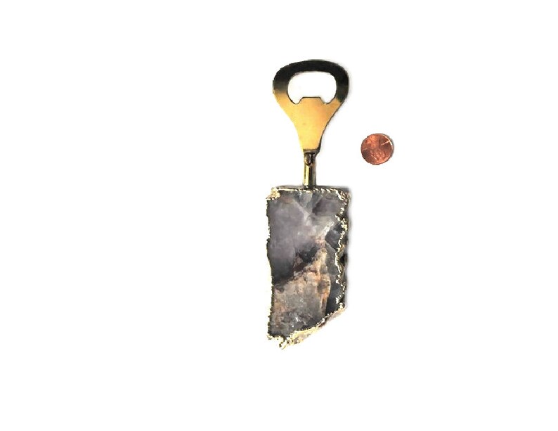 May include: A bottle opener with a brass finish and a natural stone handle. The stone is a mix of gray and purple with gold edging. A penny is next to the bottle opener for size comparison.