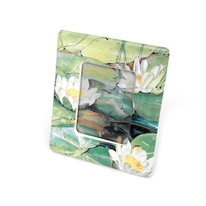 May include: A square picture frame featuring a painted design of white water lilies with yellow centers surrounded by green lily pads. The frame has a glossy finish and a central rectangular opening for displaying a photograph or artwork.