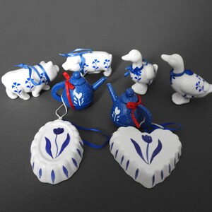 May include: A collection of ceramic ornaments featuring a blue and white floral design. The set includes pig and duck figurines, teapots, and oval and heart-shaped ornaments. Each piece is adorned with blue ribbons or accents.