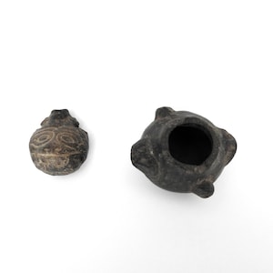 May include: Two small, dark gray, ancient-looking pottery pieces. One is a small, round vessel with a circular opening. The other is a small, rounded object with a carved spiral design.
