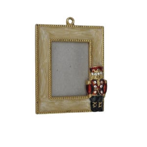 May include: A rectangular picture frame with a cream-colored, marbled enamel finish and a beaded gold border. A small, red, black, and gold nutcracker figure embellished with clear rhinestones is attached to the right side of the frame.