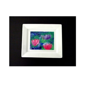 May include: A white rectangular ceramic dish with a painted floral design. The artwork features three flowers in shades of pink and purple against a green and blue background. The dish is set against a black background.