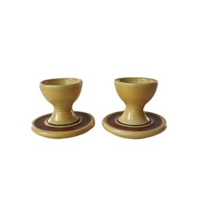 May include: Two ceramic egg cups with a beige finish. Each cup has a rounded bowl shape, a pedestal base, and a circular base with a brown and black accent. The egg cups are designed to hold eggs.
