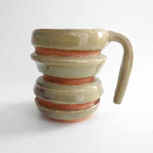 May include: A handmade ceramic mug with a unique, stacked design. The mug features a light green glaze with bands of orange-red clay. The handle is curved and the mug is approximately 4 inches tall.