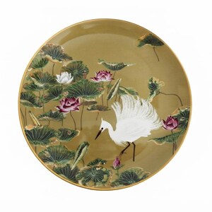 May include: A round decorative plate with a gold-toned rim featuring a detailed illustration of a white egret standing among green lotus leaves and blooming pink and white lotus flowers against a muted golden-brown background.