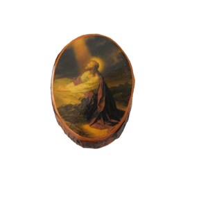 May include: An oval-shaped wooden plaque featuring a printed image of a figure with a glowing halo, kneeling in prayer. The figure wears a light-colored tunic and a dark robe, positioned against a dark, atmospheric background with a beam of light.