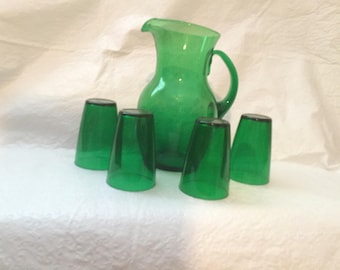 May include: A green glass pitcher with a handle and spout, accompanied by five matching green glasses. The glassware set is displayed on a white surface, creating a striking contrast with the vibrant green color.