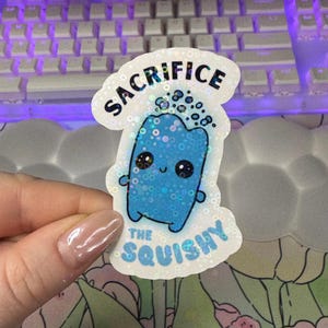 May include: A white sticker with blue glitter features a blue cartoon character with the text "Sacrifice the squishy" in black.