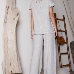 May include: A light gray linen pantsuit with a relaxed fit. The top has a round neckline and short sleeves with ruffles. The pants are wide-legged and have a straight fit.