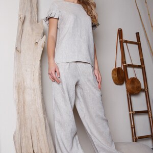 May include: A woman wearing a light beige linen top with ruffled sleeves and matching wide-leg pants. She is standing next to a large piece of driftwood and a bamboo ladder.