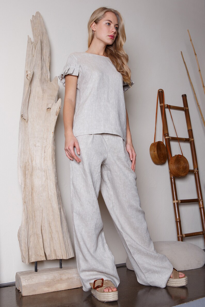 May include: A woman wearing a light beige linen top with ruffled sleeves and matching wide-leg pants. She is standing next to a large piece of driftwood and a bamboo ladder.