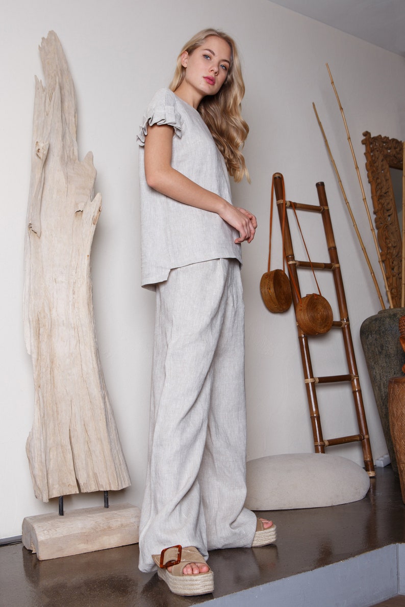 May include: A woman wearing a light gray linen top and matching wide-leg pants. She is standing in front of a wooden sculpture and a bamboo ladder with two round woven bags hanging from it.