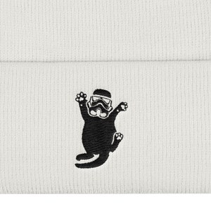 May include: A white knit beanie featuring a black embroidered graphic of a cat wearing a stormtrooper helmet. The cat is depicted in a jumping or pouncing pose with its front paws raised and its tail curved, centered on the folded cuff.