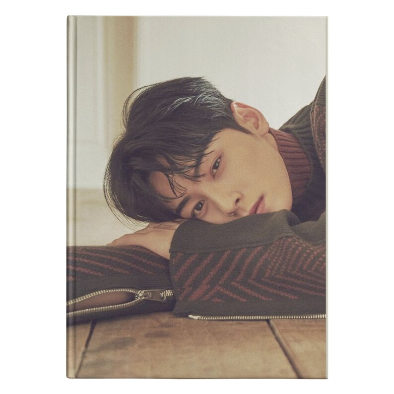May include: A young man with dark hair is lying on his side, resting his head on his hand. He is wearing a brown and red patterned sweater. The background is a light beige.
