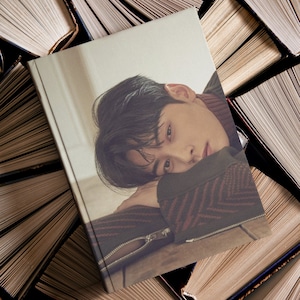 May include: A photo book cover featuring a young man with dark hair, wearing a red and black patterned sweater, resting his head on his hand. The background is a stack of books.