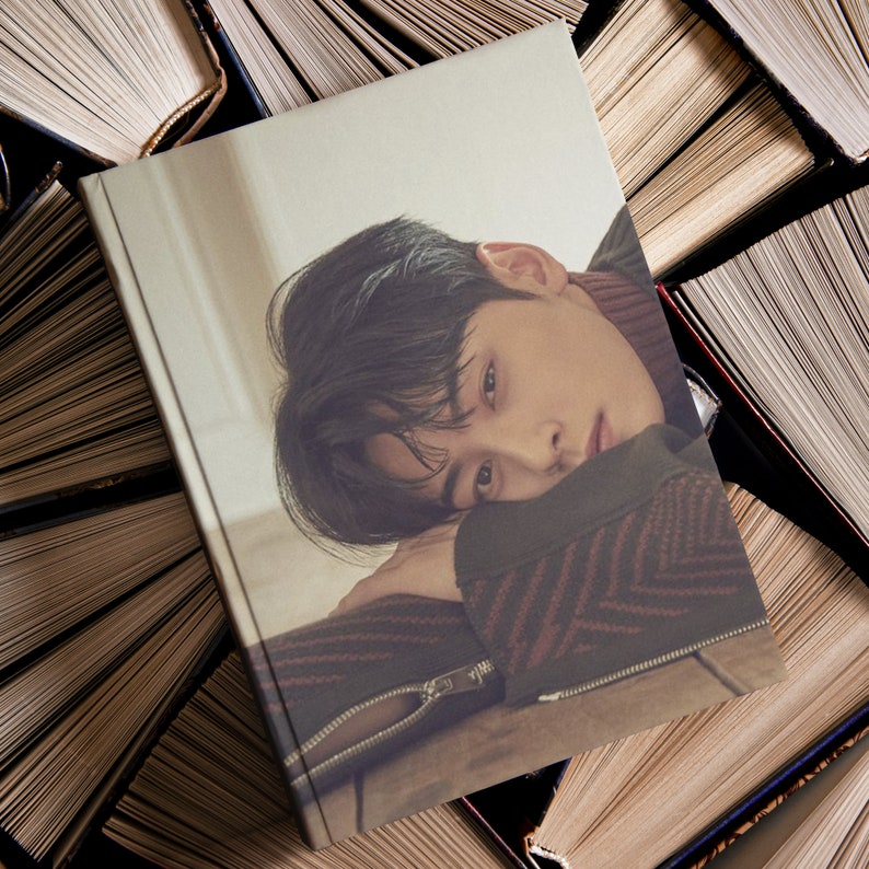 May include: A photo book cover featuring a young man with dark hair, wearing a red and black patterned sweater, resting his head on his hand. The background is a stack of books.