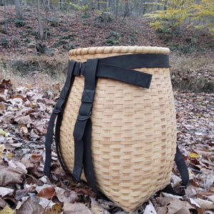 May include: A light brown, woven basket backpack with black straps and buckles. The basket is cylindrical with a wide opening and is sitting on a bed of fallen leaves in a forest setting.