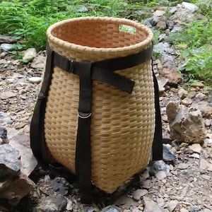May include: A tall, cylindrical woven basket made of light-colored wood strips. The basket features a wide, dark brown fabric strap that encircles the upper rim and extends downward, secured with metal D-rings on the exterior of the woven body.