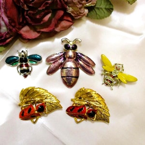 May include: A collection of insect-themed brooches. The brooches include a green and red bee, a pink and purple bee, a yellow bee, and two leaf-shaped brooches with red ladybugs. The brooches are made of metal and enamel.
