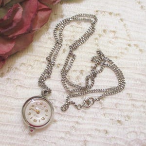 May include: A silver-coloured necklace featuring a round watch pendant. The watch face is white with gold hands and numerals. The chain is a delicate curb link style, and the watch has a small red accent.