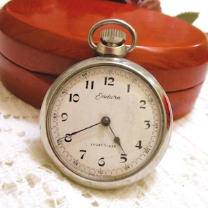 May include: A silver Endura Sport Timer pocket watch with a white face and black numerals. The watch has a silver crown and hands, and the words "Sport Timer" are printed on the face. A red, heart-shaped box is in the background.