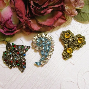 May include: Three vintage brooches are displayed on a white surface, with a backdrop of dried roses. The brooches include a leaf design with colorful rhinestones, a blue rhinestone brooch, and a gold-toned brooch with yellow stones.