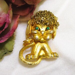 May include: A vintage gold-tone brooch in the shape of a seated poodle. The poodle has green rhinestone eyes and a chain-link textured head. The brooch is detailed with a curled tail and is set against a white background.