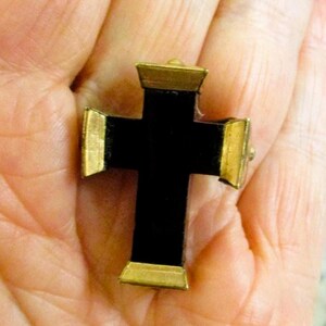 Puede incluir: Un peque&ntilde;o colgante de cruz con un cuerpo central negro mate y tapas met&aacute;licas de tono dorado en cada uno de los cuatro extremos. La cruz tiene un dise&ntilde;o geom&eacute;trico estructurado con superficies planas y bordes definidos.