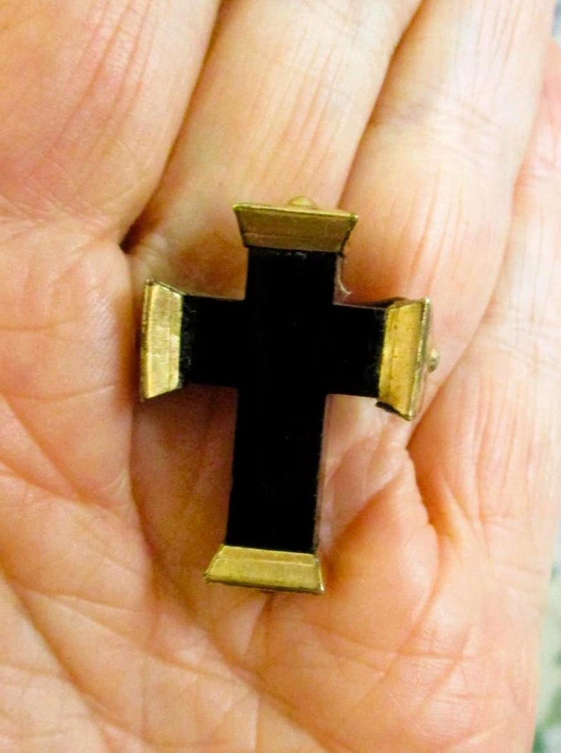 Puede incluir: Un peque&ntilde;o colgante de cruz con un cuerpo central negro mate y tapas met&aacute;licas de tono dorado en cada uno de los cuatro extremos. La cruz tiene un dise&ntilde;o geom&eacute;trico estructurado con superficies planas y bordes definidos.
