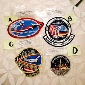 May include: Four vintage space shuttle mission patches. The patches feature colourful designs with text, including "Columbia," "Spacelab," and names of astronauts. Each patch is in a plastic sleeve.