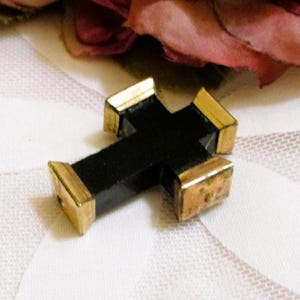 Puede incluir: Un colgante de cruz negra con tapas de metal en tono dorado pulido en cada uno de los cuatro extremos. La cruz tiene una estructura central negra mate y lisa con forma geom&eacute;trica, que contrasta con las piezas finales de metal dorado.