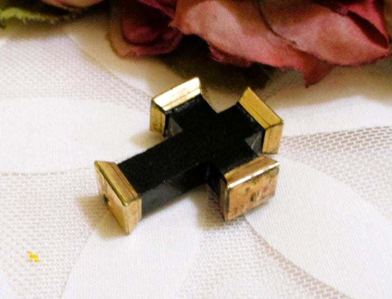 Puede incluir: Un colgante de cruz negra con tapas de metal en tono dorado pulido en cada uno de los cuatro extremos. La cruz tiene una estructura central negra mate y lisa con forma geom&eacute;trica, que contrasta con las piezas finales de metal dorado.