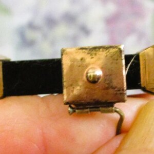 Puede incluir: Un broche rectangular de metal dorado con una placa cuadrada central que tiene un peque&ntilde;o punto en relieve, flanqueado por dos piezas finales trapezoidales m&aacute;s peque&ntilde;as, unidas por una banda horizontal negra con un mecanismo de bisagra.