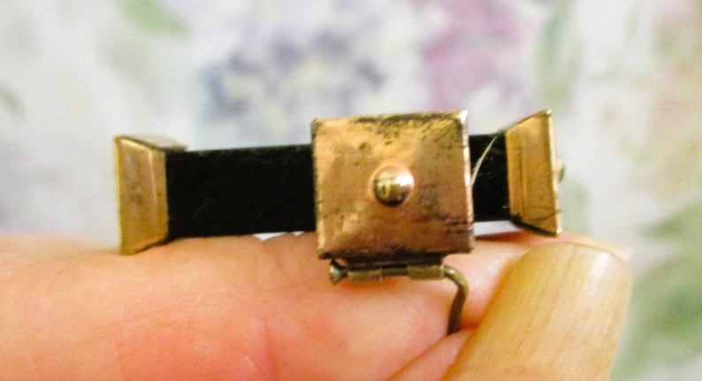 Puede incluir: Un broche rectangular de metal dorado con una placa cuadrada central que tiene un peque&ntilde;o punto en relieve, flanqueado por dos piezas finales trapezoidales m&aacute;s peque&ntilde;as, unidas por una banda horizontal negra con un mecanismo de bisagra.