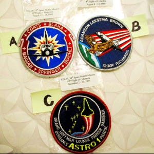 May include: Three vintage space shuttle mission patches. Patch A features a shuttle and starburst design with the names of the crew. Patch B shows an eagle and shuttle over a red and white striped background. Patch C has a rocket and the word "ASTRO".