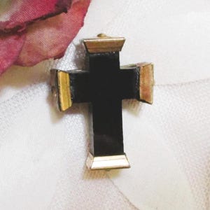 Puede incluir: Un colgante de cruz con un cuerpo central negro liso y tapas met&aacute;licas en tono dorado en cada uno de los cuatro extremos. La cruz presenta un dise&ntilde;o geom&eacute;trico estructurado con l&iacute;neas limpias y un acabado pulido, creando un marcado contraste entre el centro oscuro y los bordes reflectantes de color dorado.
