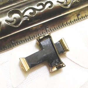 Puede incluir: Un broche en forma de cruz negra con extremos de metal dorado y un mecanismo de alfiler en la parte posterior. El broche est&aacute; colocado junto a una regla de metal, mostrando que el art&iacute;culo mide aproximadamente 3,8 cm de ancho y 3,2 cm de alto.