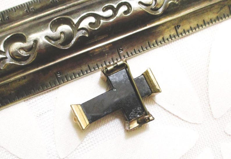 Puede incluir: Un broche en forma de cruz negra con extremos de metal dorado y un mecanismo de alfiler en la parte posterior. El broche est&aacute; colocado junto a una regla de metal, mostrando que el art&iacute;culo mide aproximadamente 3,8 cm de ancho y 3,2 cm de alto.