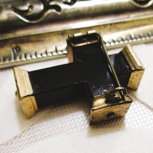 Puede incluir: Un broche en forma de cruz negra con extremos de metal dorado y un mecanismo de alfiler en la parte posterior. Una regla de metal es visible al fondo, mostrando que el objeto mide aproximadamente 3,8 cm de ancho en la barra horizontal.