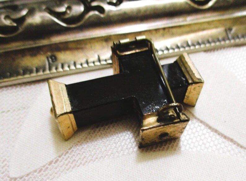 Puede incluir: Un broche en forma de cruz negra con extremos de metal dorado y un mecanismo de alfiler en la parte posterior. Una regla de metal es visible al fondo, mostrando que el objeto mide aproximadamente 3,8 cm de ancho en la barra horizontal.