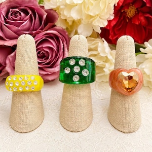 May include: Three chunky rings displayed on beige fabric cones. The left ring is bright yellow with embedded clear rhinestones. The center ring is translucent green with clear rhinestones. The right ring is peach-colored with a large heart-shaped gem.