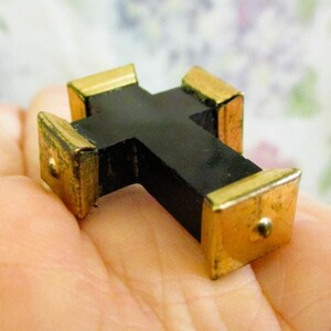 Puede incluir: Un peque&ntilde;o objeto en forma de cruz con una estructura central negra mate y tapas de metal dorado pulido en cada uno de los cuatro extremos. Cada tapa dorada tiene forma cuadrada y presenta un peque&ntilde;o detalle de remache met&aacute;lico centrado.