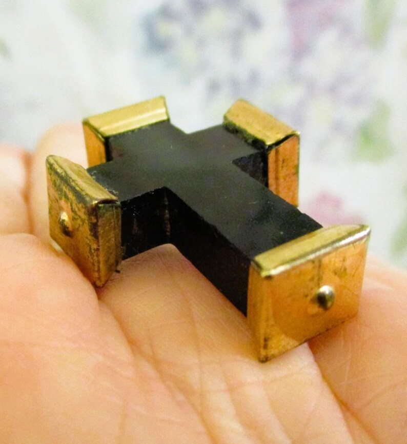 Puede incluir: Un peque&ntilde;o objeto en forma de cruz con una estructura central negra mate y tapas de metal dorado pulido en cada uno de los cuatro extremos. Cada tapa dorada tiene forma cuadrada y presenta un peque&ntilde;o detalle de remache met&aacute;lico centrado.