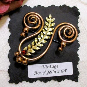 May include: Vintage rose and yellow gold-tone brooch with a leaf and swirl design. The brooch features a small red gemstone and several gold-tone spheres. The text "Vintage Rose/Yellow GF" is printed on a white label.