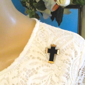 Puede incluir: Un broche en forma de cruz con un centro negro s&oacute;lido y bordes met&aacute;licos en tono dorado en la parte superior, inferior y los lados. El broche est&aacute; prendido en una prenda de encaje blanco con un patr&oacute;n floral texturizado y cuello redondo.