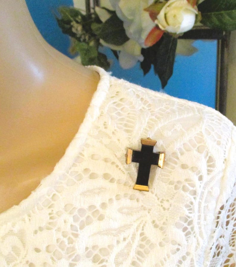 Puede incluir: Un broche en forma de cruz con un centro negro s&oacute;lido y bordes met&aacute;licos en tono dorado en la parte superior, inferior y los lados. El broche est&aacute; prendido en una prenda de encaje blanco con un patr&oacute;n floral texturizado y cuello redondo.