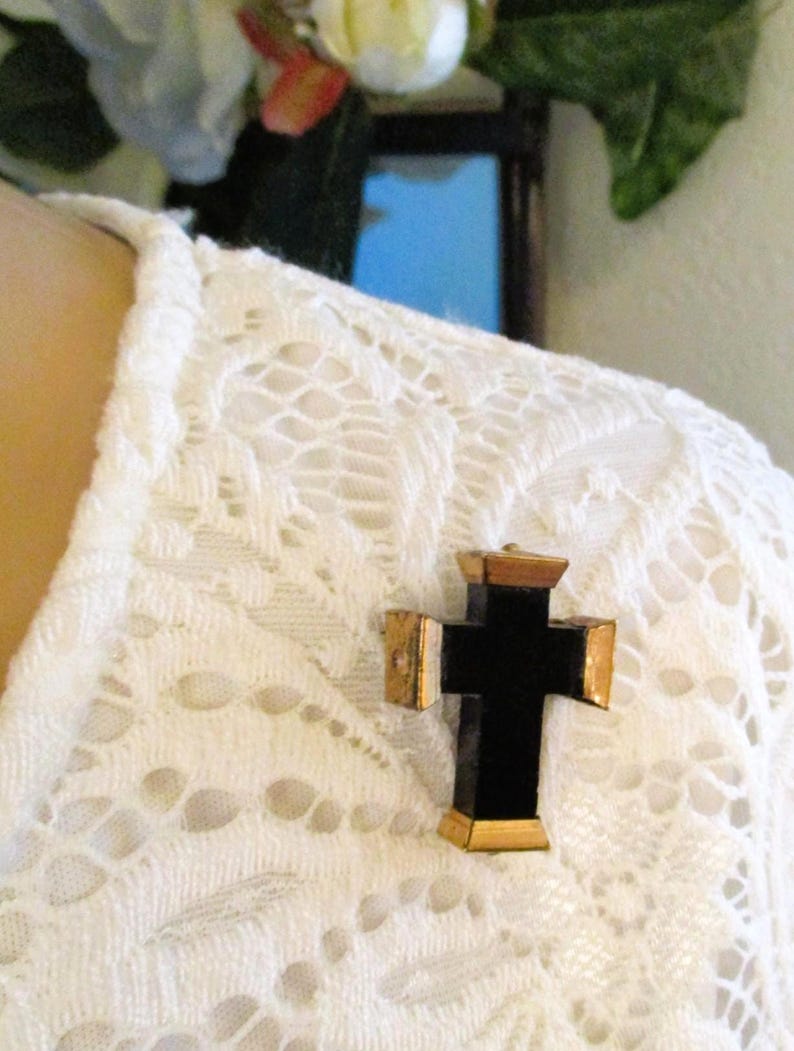 Puede incluir: Un broche en forma de cruz con un cuerpo central negro liso y tapas met&aacute;licas en tono dorado en cada uno de los cuatro extremos. La cruz est&aacute; prendida en una prenda blanca con un patr&oacute;n texturizado e intrincado similar al encaje.