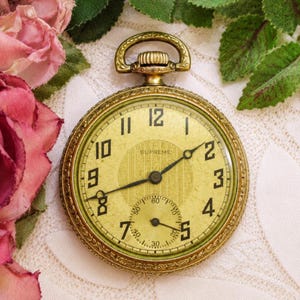 May include: A gold-tone pocket watch with an ornate, engraved circular case and a bow-shaped crown. The watch face is a pale yellow with black Arabic numerals, a central textured pattern, and a small sub-dial for seconds at the bottom.