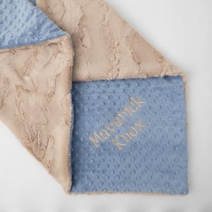 May include: A soft, two-sided blanket featuring a beige faux fur side with a textured, wavy pattern and a light blue minky side with a raised dot pattern. The blue side is embroidered with the name Maverick Knox in gold-colored thread.