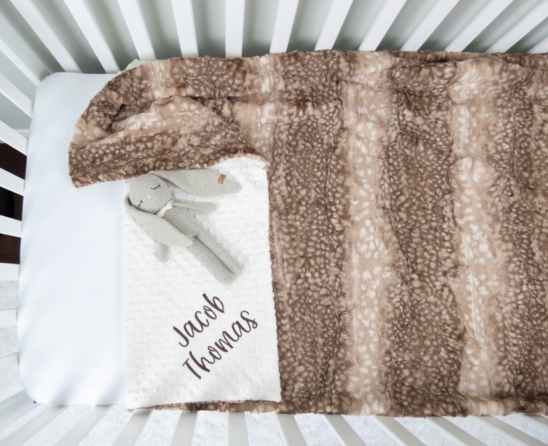 May include: A white hooded baby blanket with a brown and white deer print on one side. The blanket is in a crib and has the name "Jacob Thomas" embroidered on it.